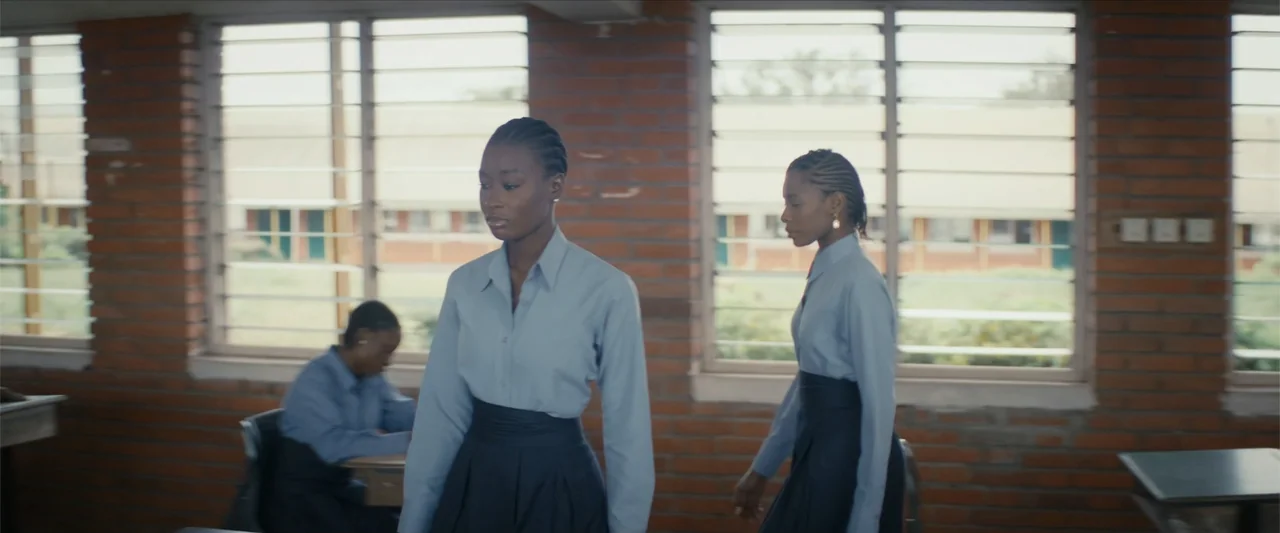 Ms. Kanyin (2025) movie still: medium — A school classroom featuring brick walls and multiple large windows overlooking an outdoor area. Two…