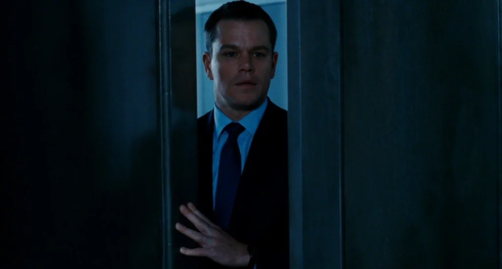 The Adjustment Bureau (2013) movie still: medium — A man stands behind a dark, partially opened heavy door in a dim environment. He reaches out with…