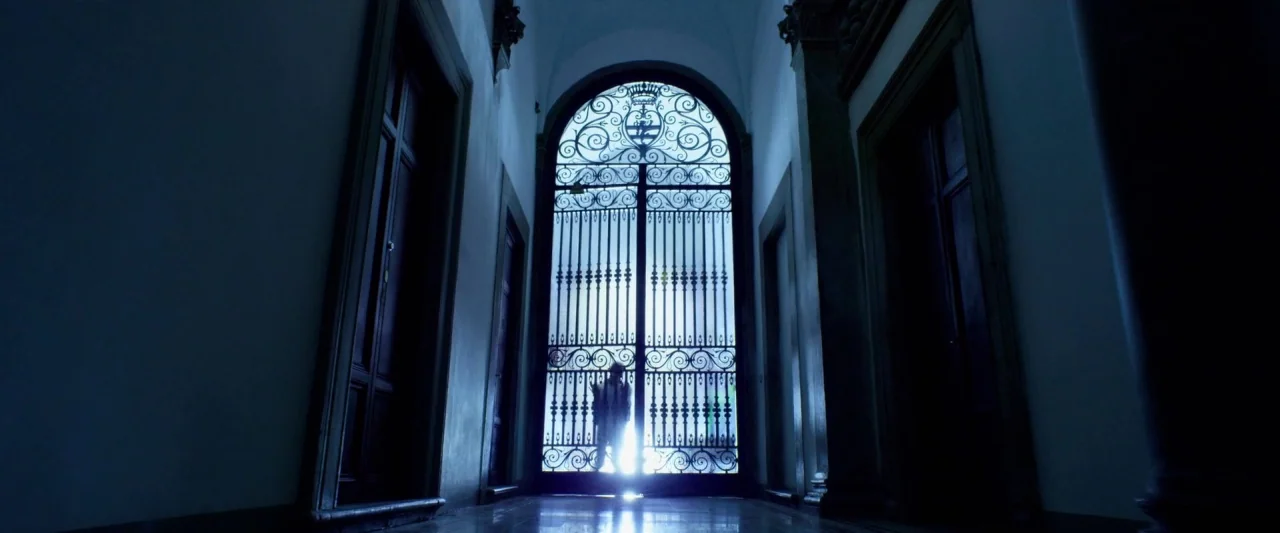 6 Underground (2019) movie still: wide — A long, dark corridor leads to an arched entrance with ornate metal gates. A lone figure stands in front of…