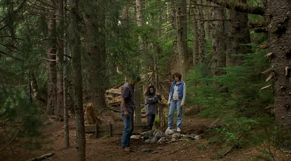 Limbo (1999) movie still: wide — A dense coniferous forest features tall trees and low undergrowth on a brown dirt floor. Three people congregate…