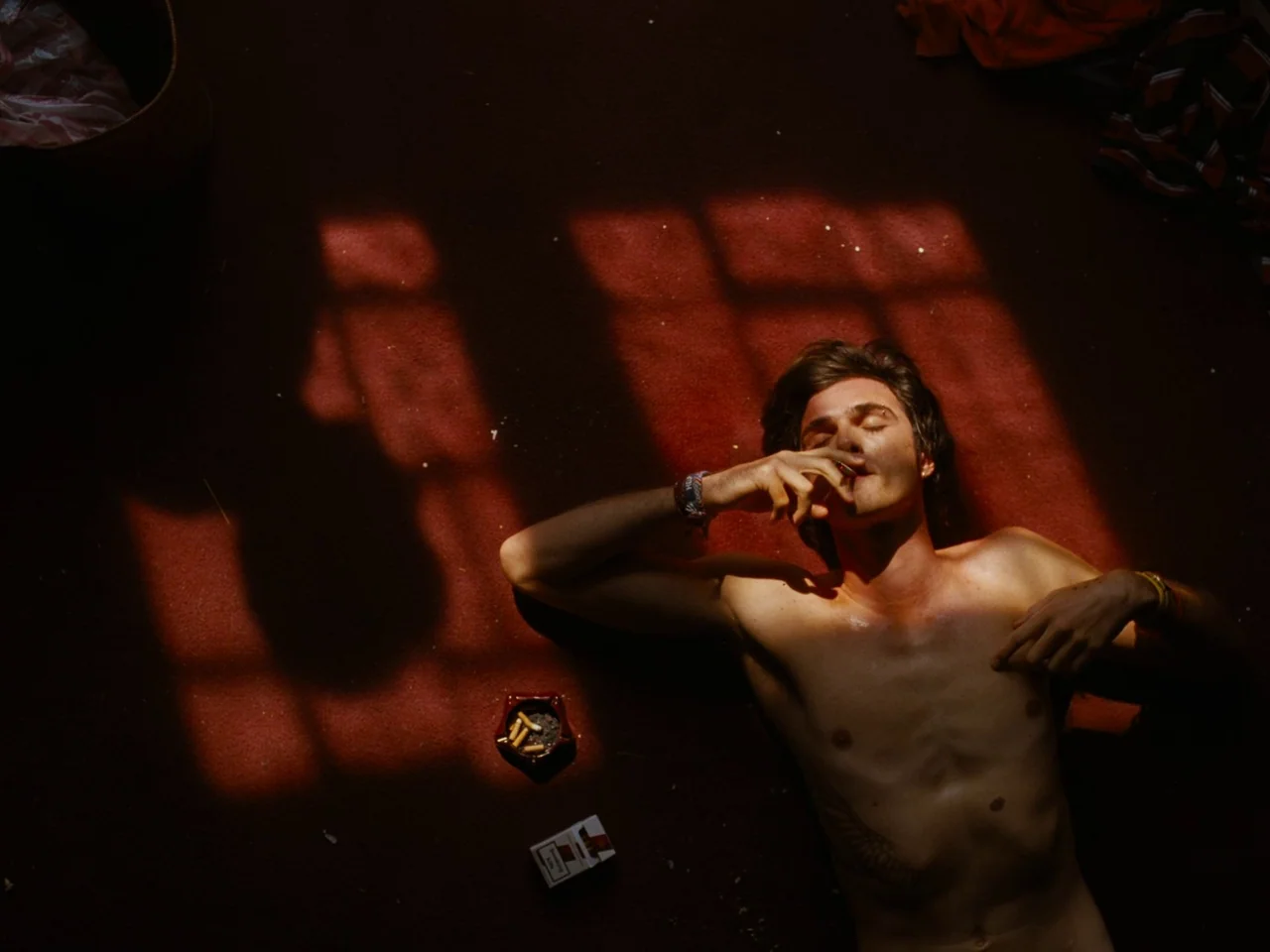 Saltburn (2023) movie still: medium — A man lies shirtless on a dark red carpeted floor with shadows projected on it. He holds a cigarette while…