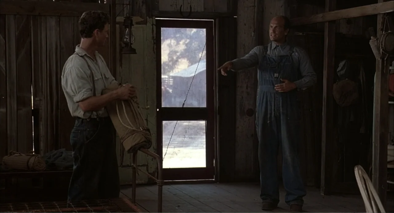 Of Mice and Men (1992) movie still: fullbody — A rustic wooden bunkhouse interior with plank walls and a screen door center frame. One man holding a…