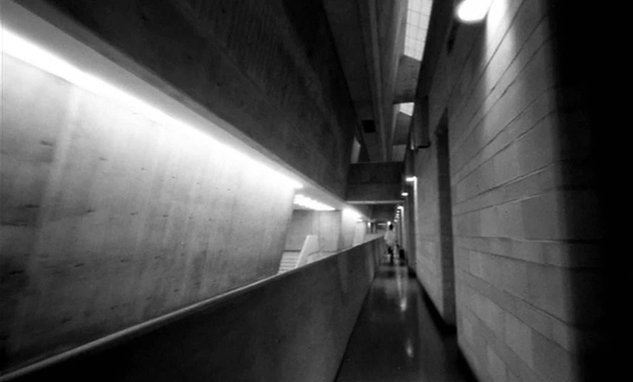 Stereo (1969) movie still: wide — A long concrete corridor features slanted walls and harsh industrial geometry. Linear light fixtures illuminate…
