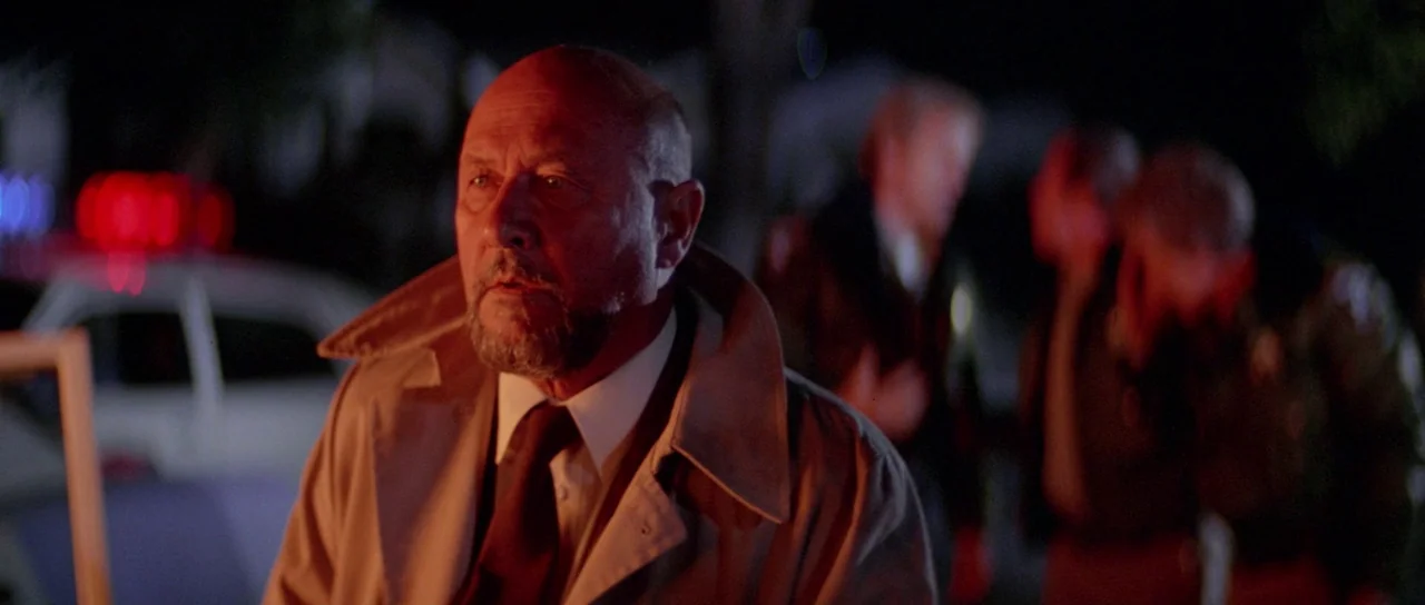 Halloween II (1981) movie still: medium — A nighttime police investigation scene with flashing red lights in the background. An older man in a tan…