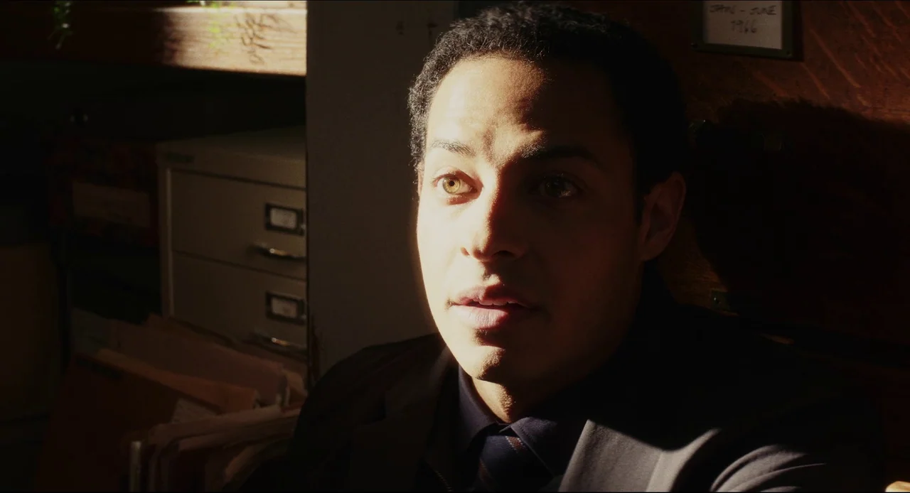 Wake Up Dead Man (2025) movie still: closeup — A man sits in a dimly lit, narrow archive room surrounded by metal filing cabinets. Strong…