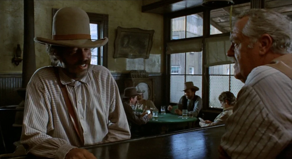 The Hired Hand (1971) movie still: medium — A wooden saloon interior with men seated at tables in the background. Two men engage in a conversation…