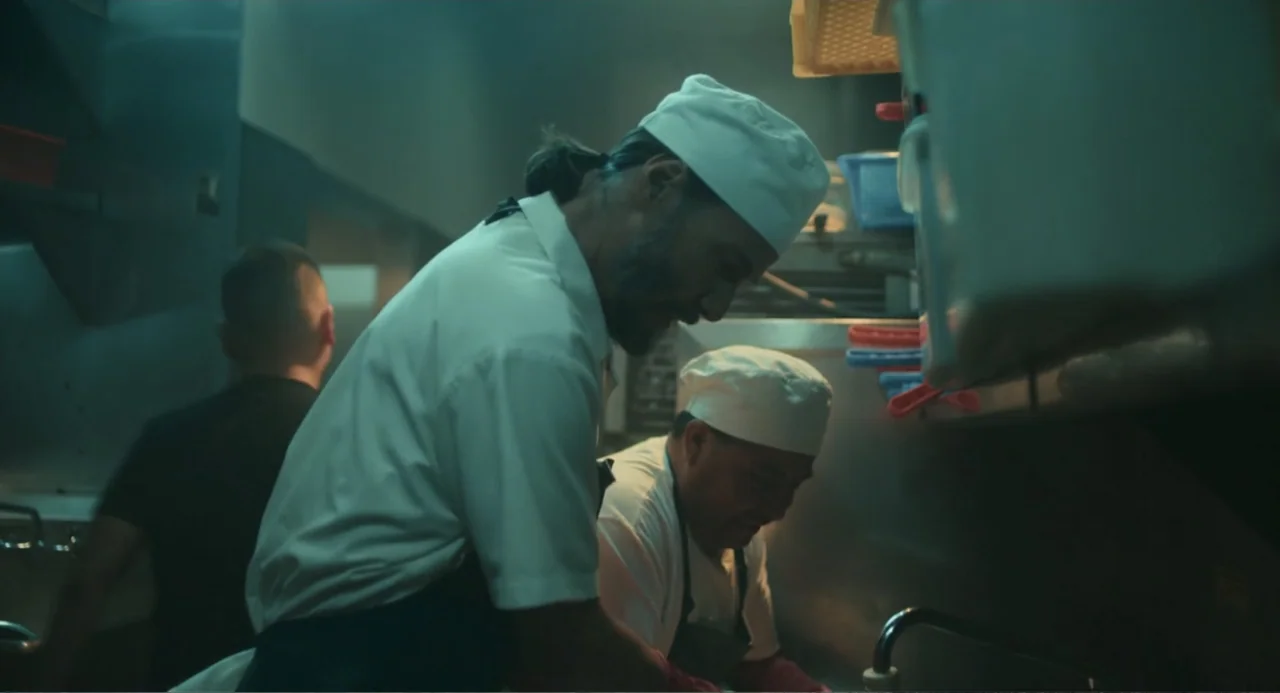 Good Fortune (2025) movie still: medium — A utilitarian restaurant kitchen with stainless steel surfaces and stacked crates. Two chefs in white…