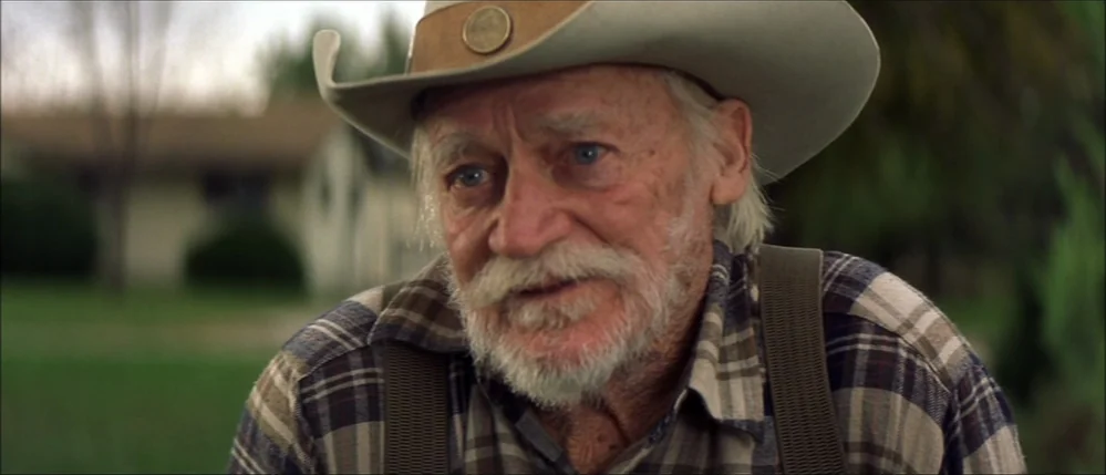 The Straight Story (1999) movie still: closeup — A man in a cowboy hat stands outdoors with a suburban house visible behind him. The man looks…