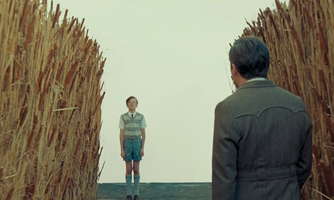 The Swan (2023) movie still: medium — Two figures stand on opposite sides of a pathway lined with tall dry reeds. A boy in school clothes faces an…