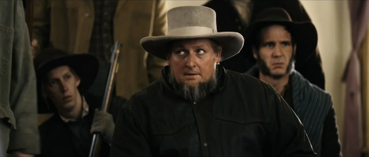 Out of Liberty (2019) movie still: medium — An interior space filled with several men in 19th-century attire. The central man in a top hat looks…