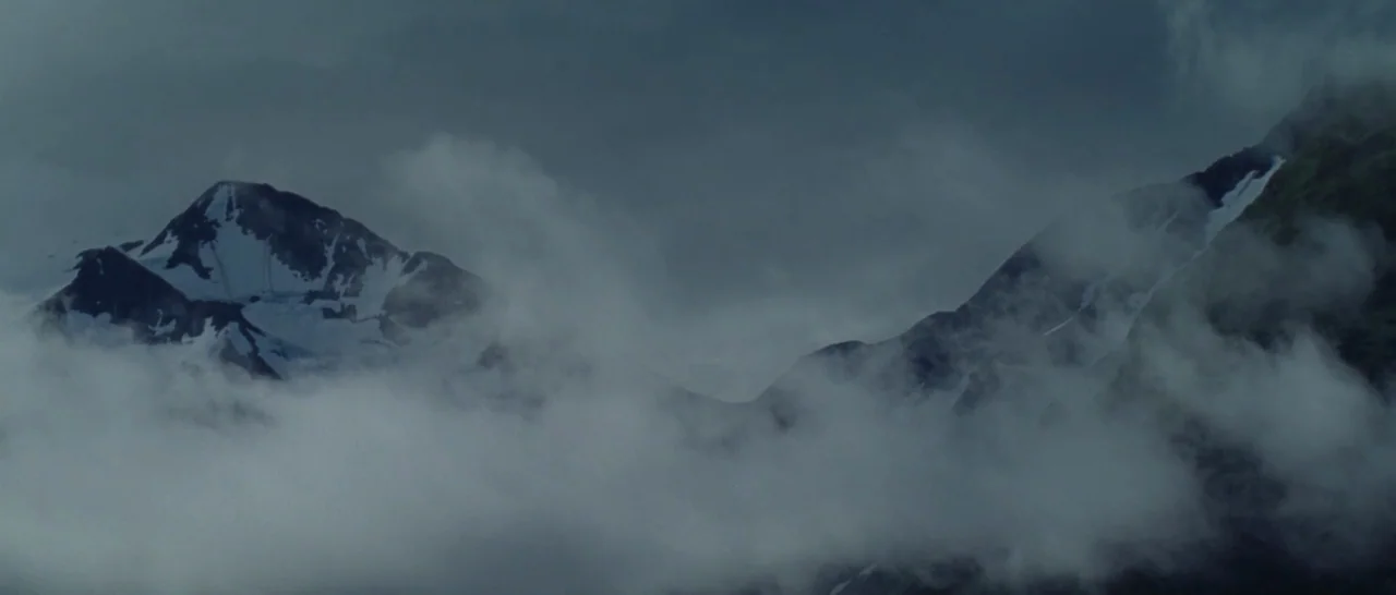 Wildlike (2014) movie still: establishing — High jagged mountain peaks with patches of snow emerge from thick, low-hanging clouds. The sky above is…