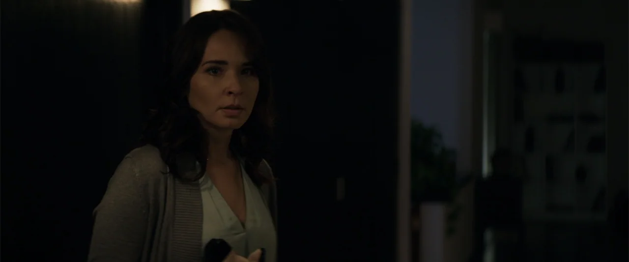 Human Animals (2020) movie still: medium — An interior space with a dimly lit background and a dark wall. A woman stands in the foreground, turning…