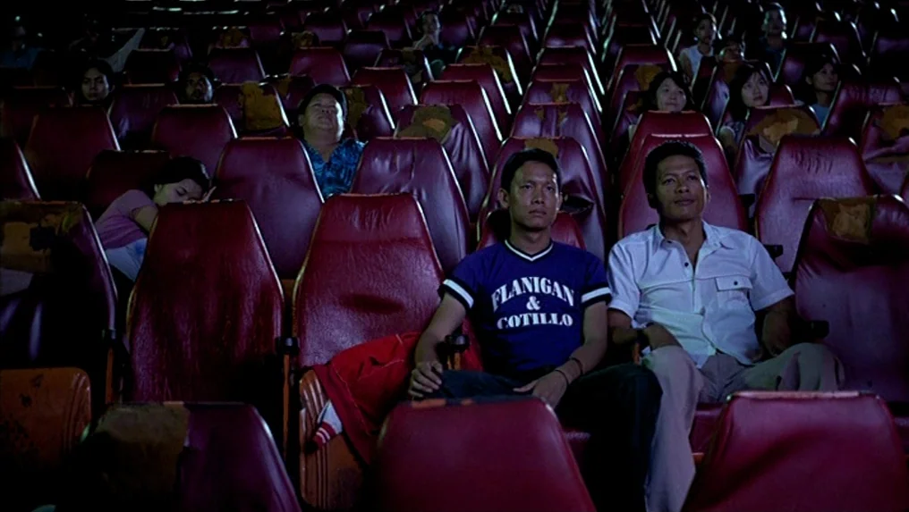 Tropical Malady (2004) movie still: medium — A dark movie theater with rows of worn red seats filled with audience members. Two men sit in the…
