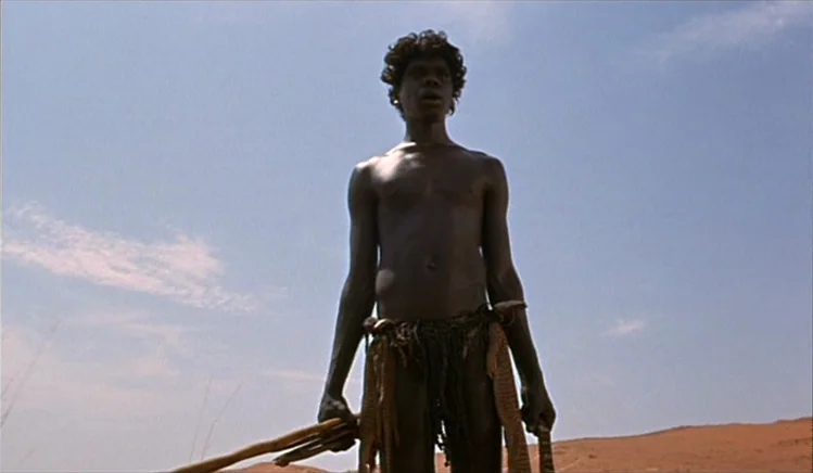 Walkabout (1971) movie still: fullbody — A vast, open desert under a pale blue sky during the daytime. A man stands upright, holding a slender…