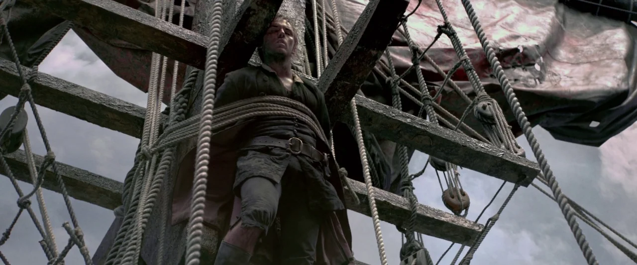 Pirates of the Caribbean: On Stranger Tides movie still: medium — A man stands tied to the wooden mast of a sailing ship with ropes. The sky is…