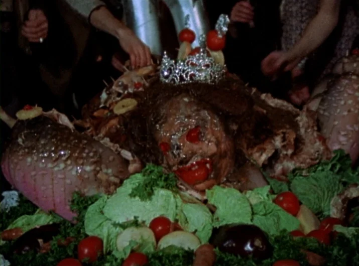 Desperate Living (1977) movie still: closeup — A centerpiece composed of cooked meats and vegetables sits on a platter. It is decorated with a small…