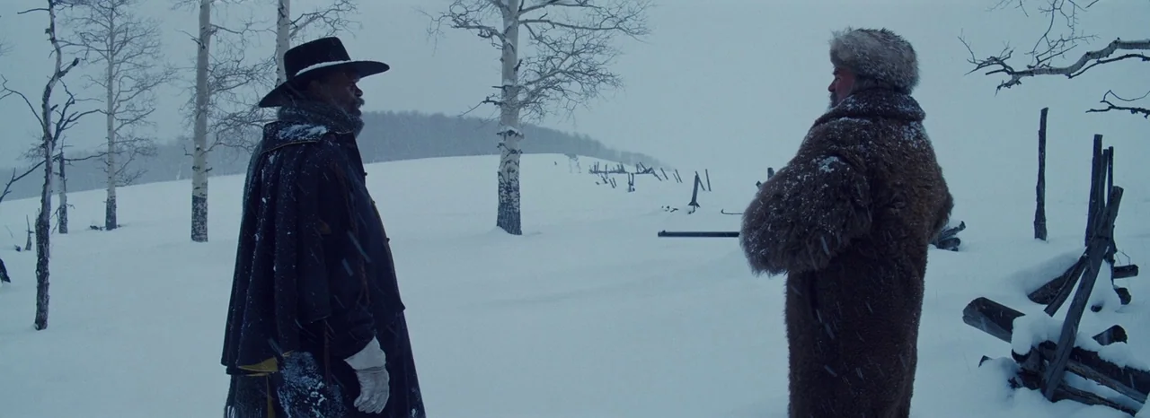The Hateful Eight (2015) movie still: fullbody — A vast, white, snow-covered landscape features sparse, bare birch trees and a rustic fence. Two men…