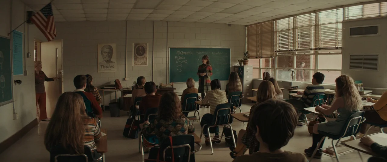 The Black Phone (2021) movie still: wide — A 1970s-era classroom with cinder block walls, a blackboard, and rows of student desks. A teacher stands…