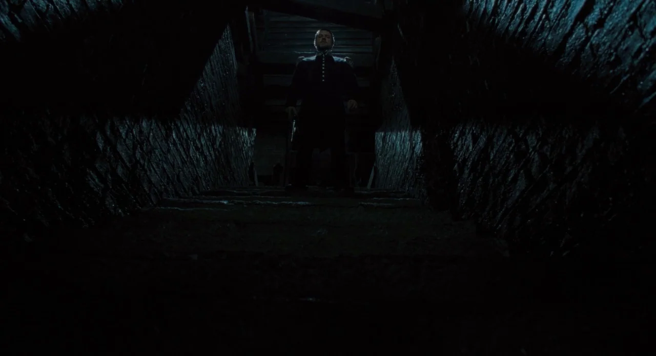 Les Misérables (2012) movie still: fullbody — A dark stone staircase is framed by rough textured brick walls in low light. A man in a dark military…