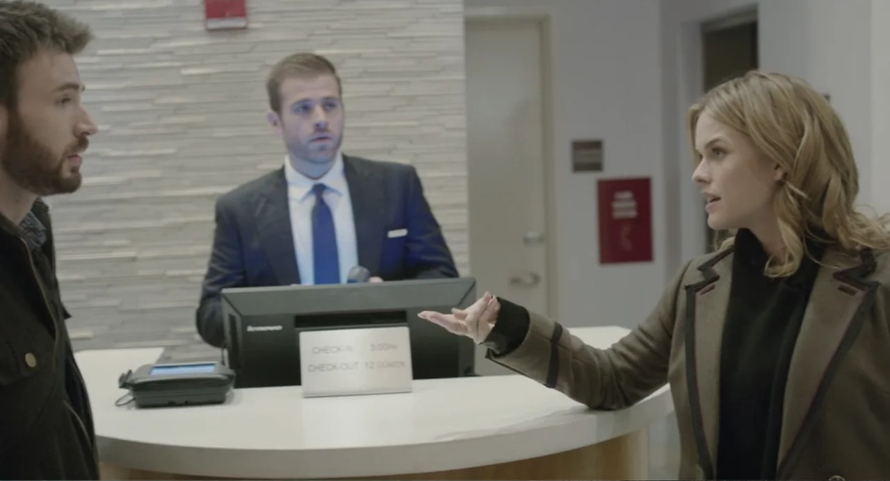 Before We Go (2014) movie still: medium — A hotel reception area features a white curved counter with a small signage placard. Two people stand in…