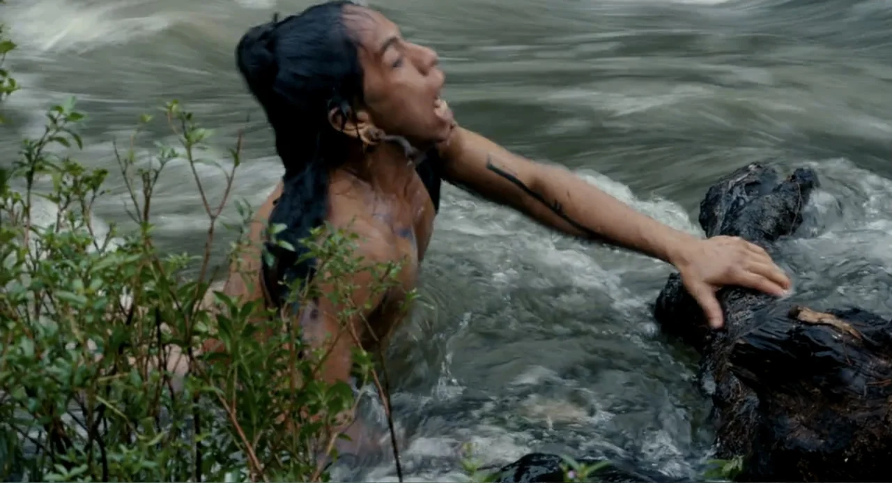 Apocalypto (2006) movie still: medium — A person struggles against the current of a shallow, rocky riverbed with greenery nearby. The shirtless man…