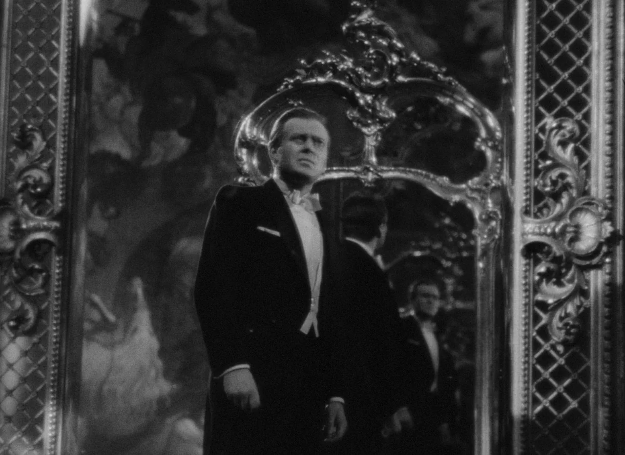 Krakatit (1948) movie still: medium — An ornate, gilded mirror frame reflects the room behind a man in a tuxedo. He stands in the foreground,…
