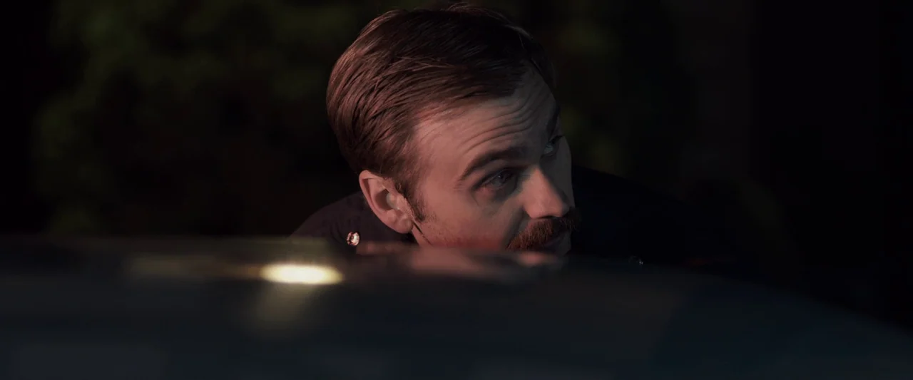 You’re Next (2013) movie still: closeup — A man peers over the dark hood of a vehicle in a nighttime setting. The focus is on the man's face as he…