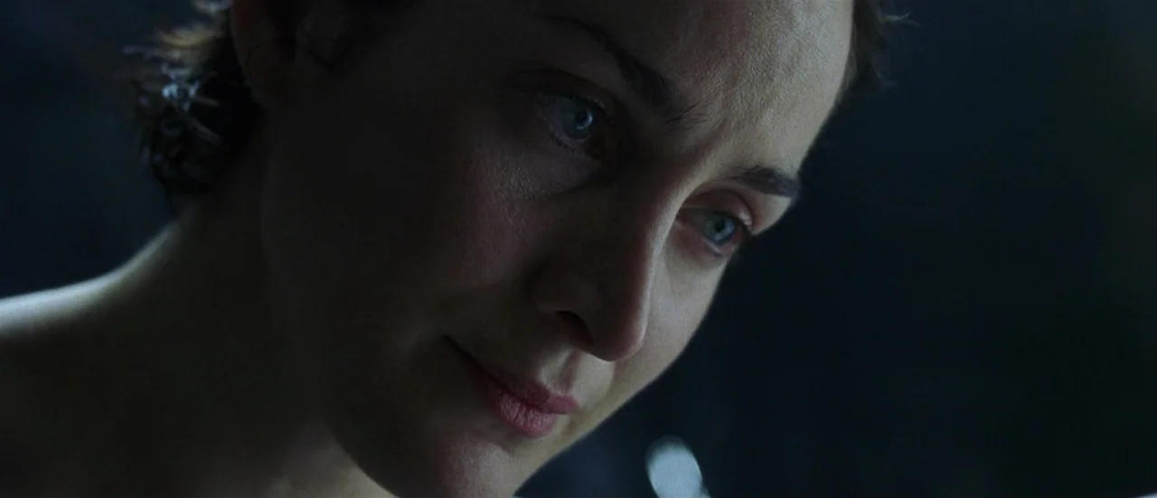The Matrix (1999) movie still: closeup — A dark, dimly lit environment provides the backdrop for a subject's face. The woman looks downwards with a…