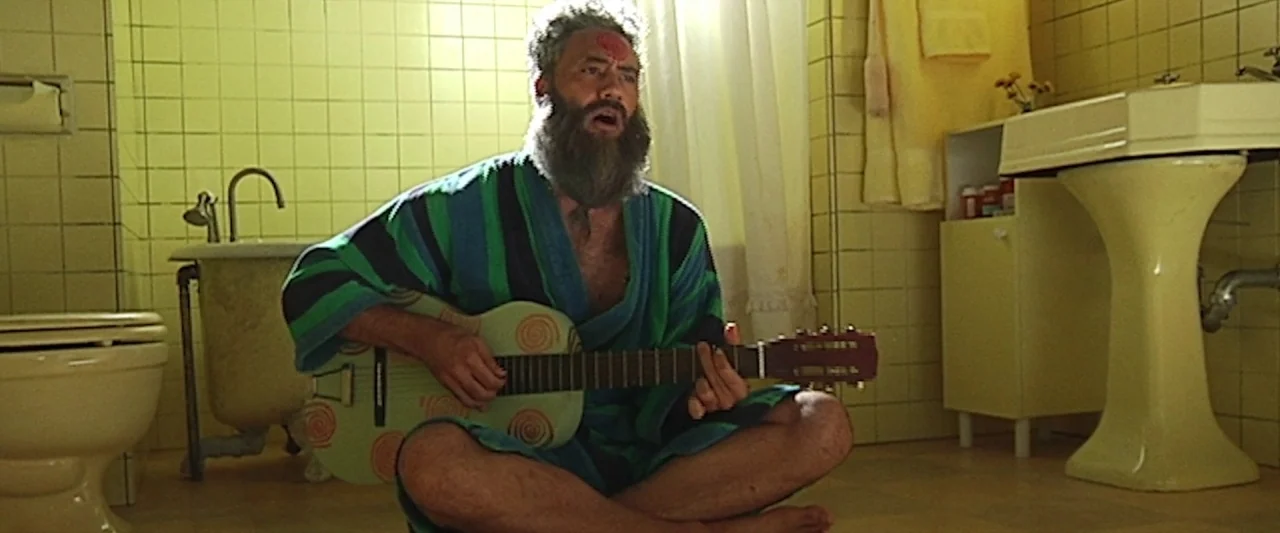 Seven Stages to Achieve Eternal Bliss (2018) movie still: medium — A yellow-tiled room features a clawfoot bathtub, pedestal sink, and a toilet. A…
