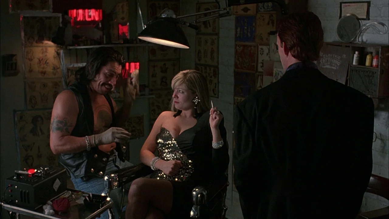 Whore (1991) movie still: medium — A dimly lit room is covered in various flash art designs on the walls. A man talks to a seated woman while a…