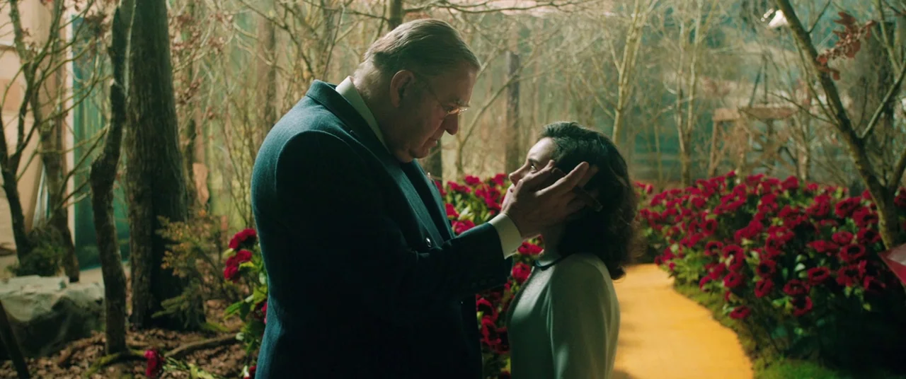 Judy (2019) movie still: medium — An interior stage set features a fake forest path with artificial red poppies. An older man stands facing a…