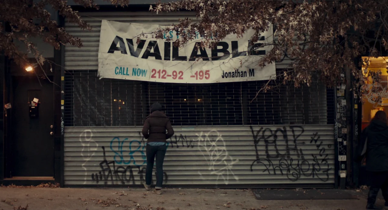 Mistress America (2015) movie still: fullbody — A city storefront has a metal security gate covered in colorful spray paint graffiti. A person…