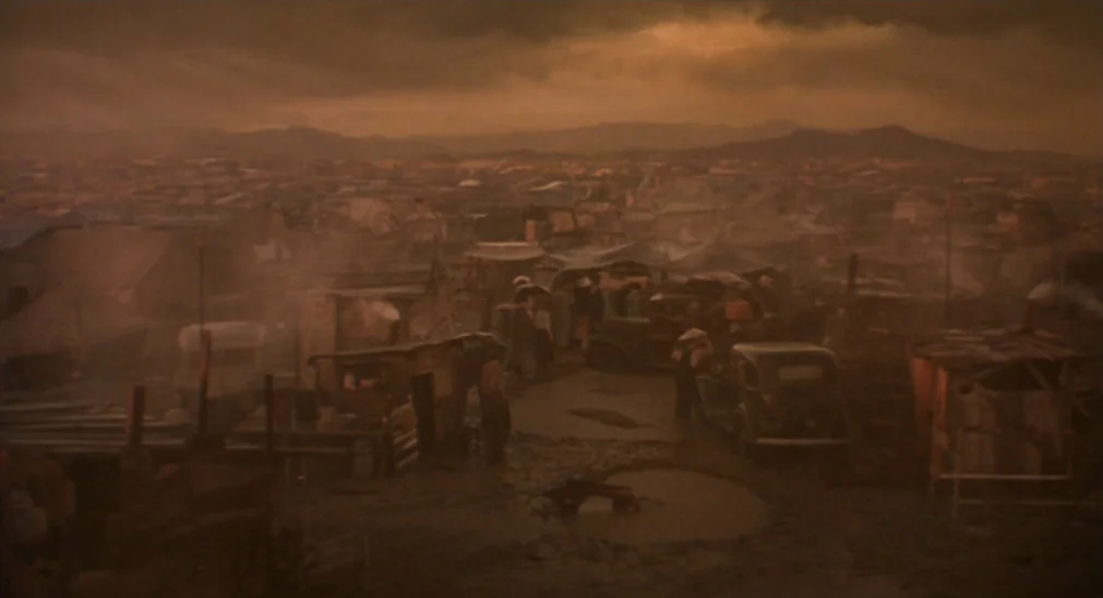 Bound For Glory (1976) movie still: establishing — A dense shantytown composed of wooden huts sits under a heavy, overcast brown sky. Dirt paths and…