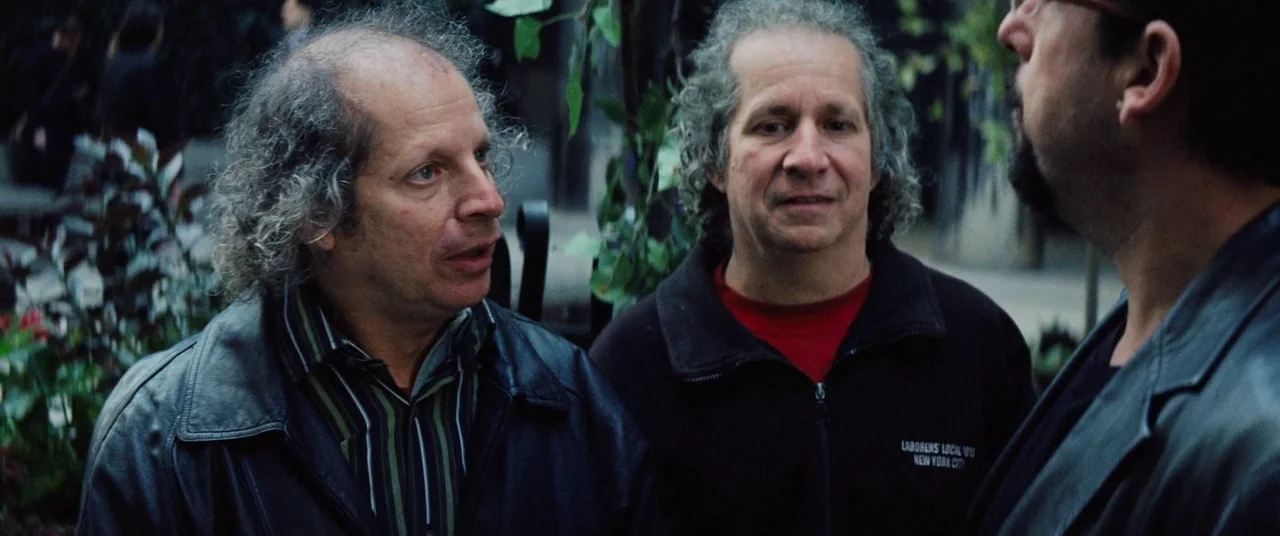 Uncut Gems (2019) movie still: medium — An urban outdoor public space features a metal railing and blurred green foliage backdrop. Three men stand…