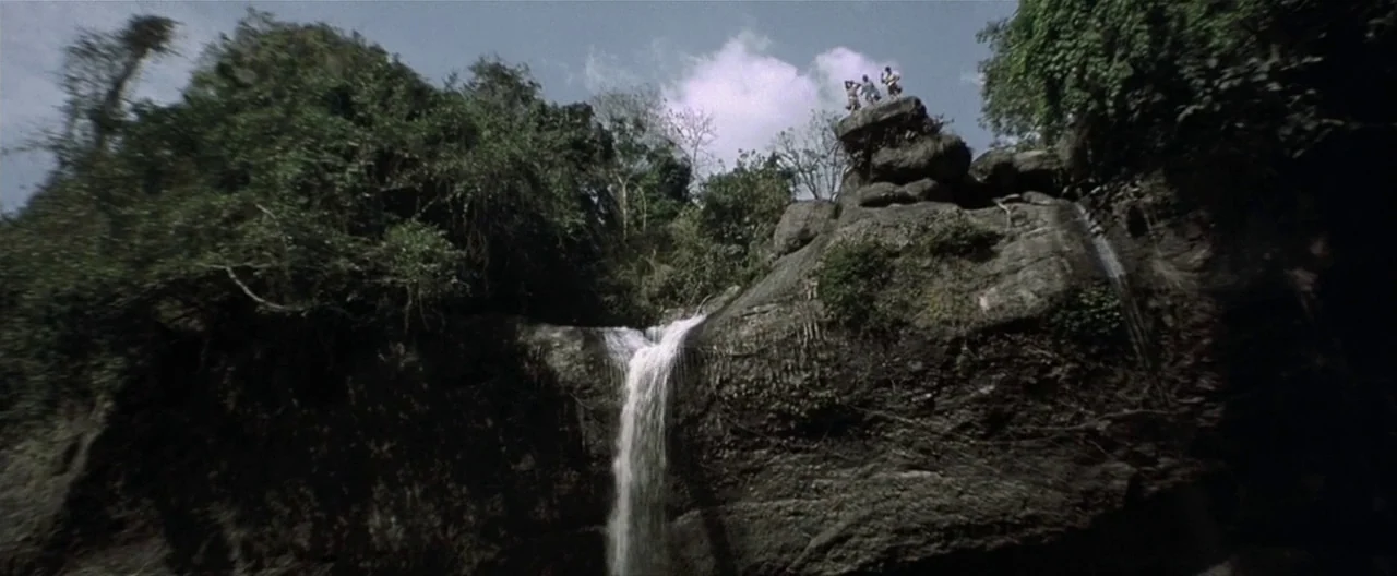 The Beach (2000) movie still: wide — A tiered rocky cliff face supports a narrow waterfall flowing into a shaded basin. Three tiny human figures…