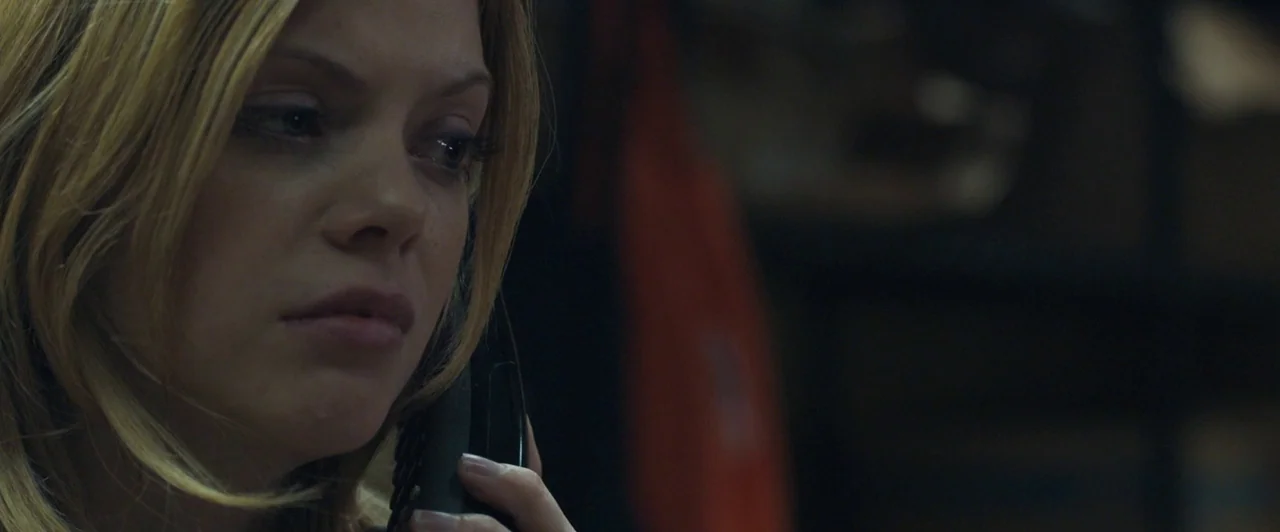 Compliance (2012) movie still: closeup — A dimly lit room provides the backdrop for a close-up of a blonde woman. She is holding a black telephone…