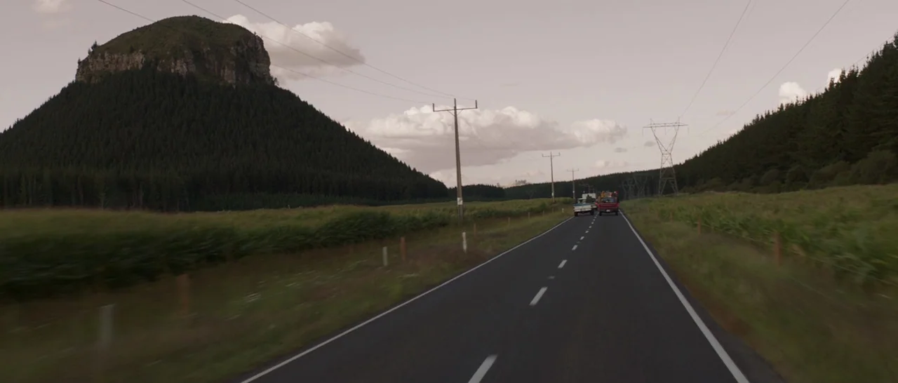 Pete’s Dragon (2016) movie still: wide — A rural asphalt road cuts through a green landscape beneath a pale sky. Two trucks drive away into the…