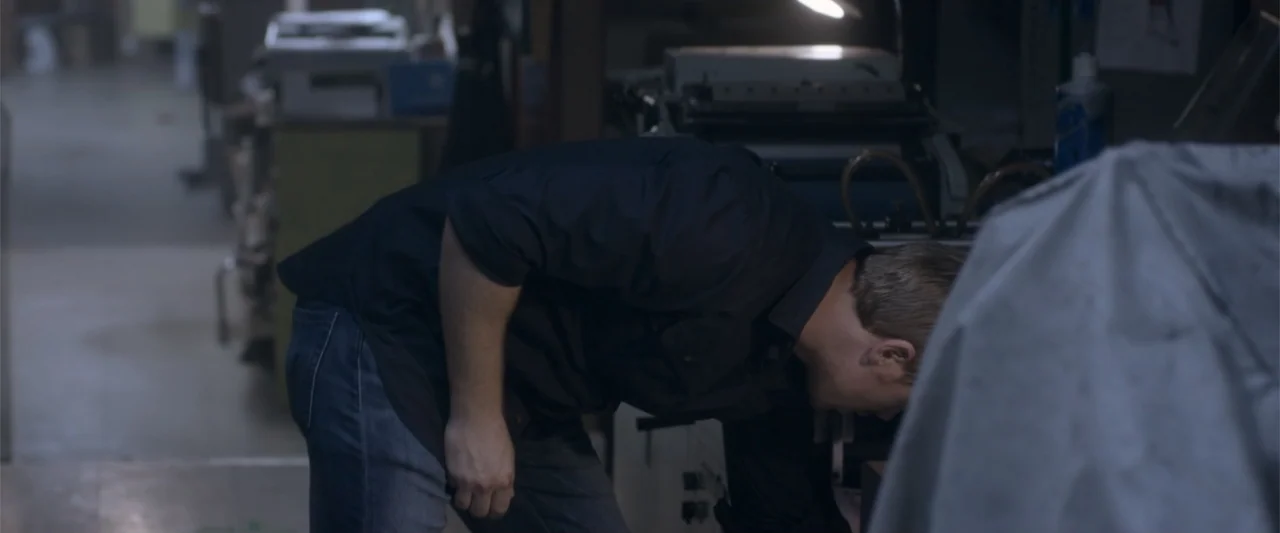 Radius (2017) movie still: medium — A cluttered printing workshop features a large industrial machine and various equipment in shadow. A man in a…