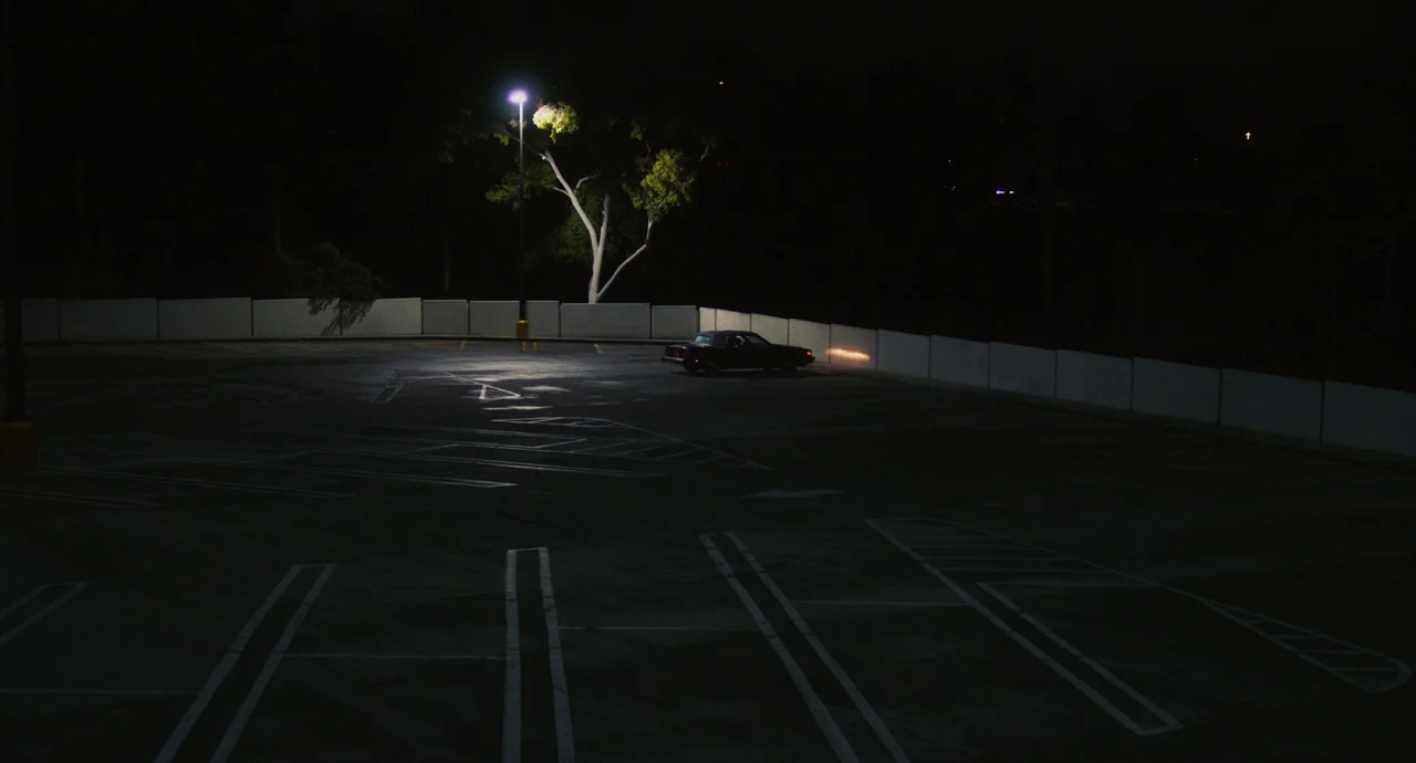 Palo Alto (2013) movie still: wide — An empty asphalt parking lot is bordered by a white concrete wall at night. A lone dark vehicle is parked…