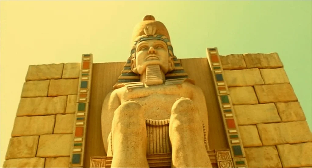 Bubba Ho-Tep (2002) movie still: fullbody — A colossal stone statue of a pharaoh sits against a brick block wall. The statue faces forward while…
