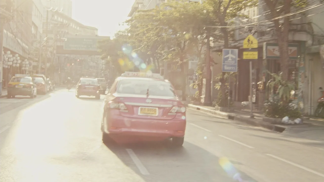 Zero Tolerance (2015) movie still: medium — A busy city street in Bangkok features a prominent pink taxi driving away. Sunlight illuminates the road…