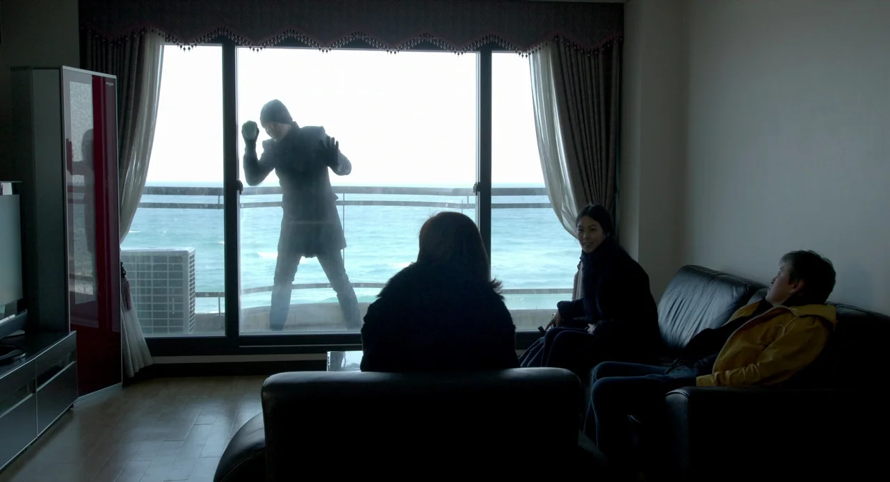 On The Beach At Night Alone (2017) movie still: wide — A modern hotel room overlooks a rough sea through a floor-to-ceiling glass wall. A hooded…