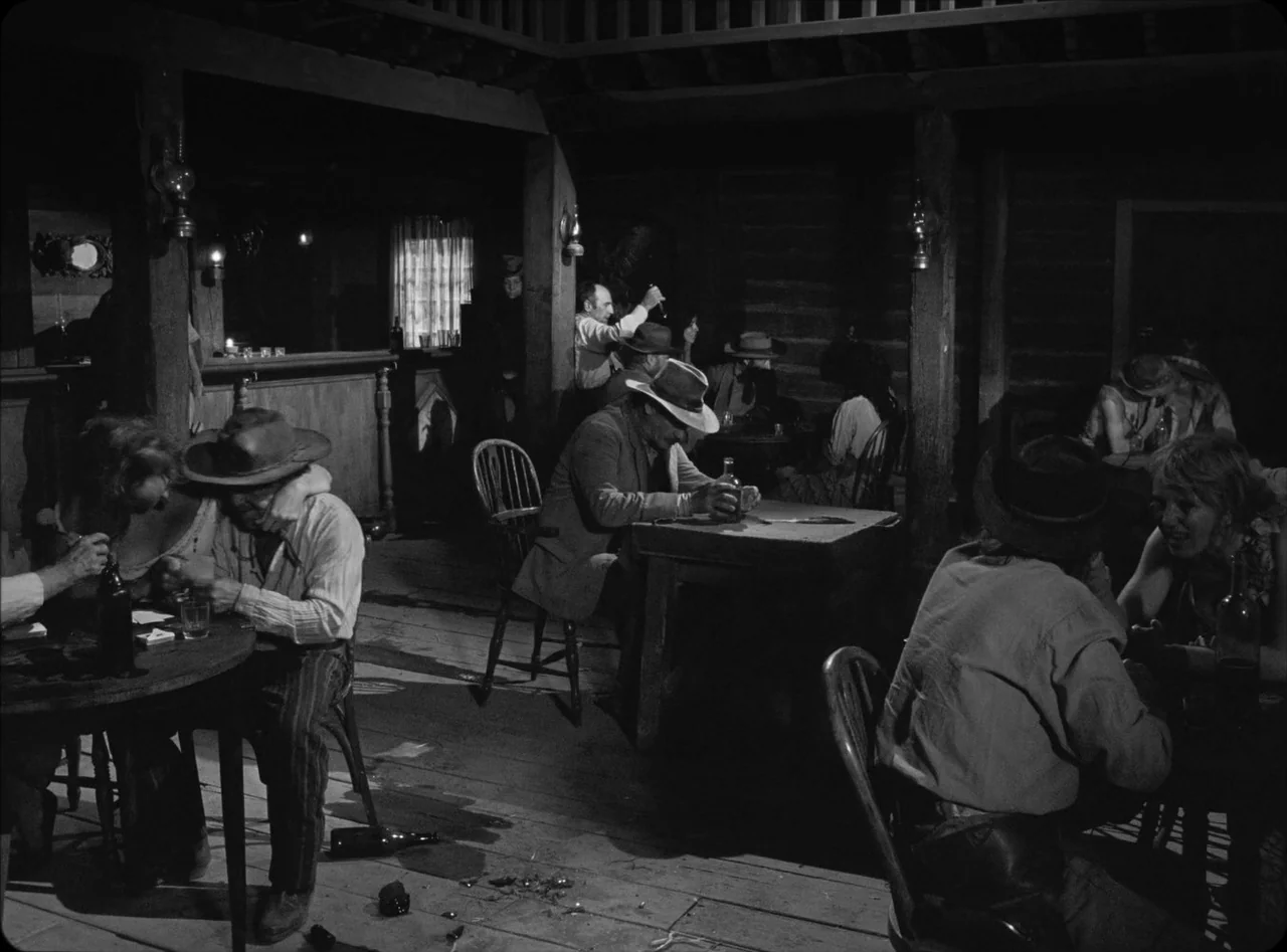 Eureka (2024) movie still: wide — A rustic saloon with wooden plank floors and simple furniture arranged for patrons. Several figures occupy the…