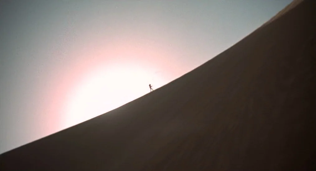 The Trip (1967) movie still: wide — A vast, empty sand dune rises against a pale sky in the desert. A lone person walks slowly up the steep incline…