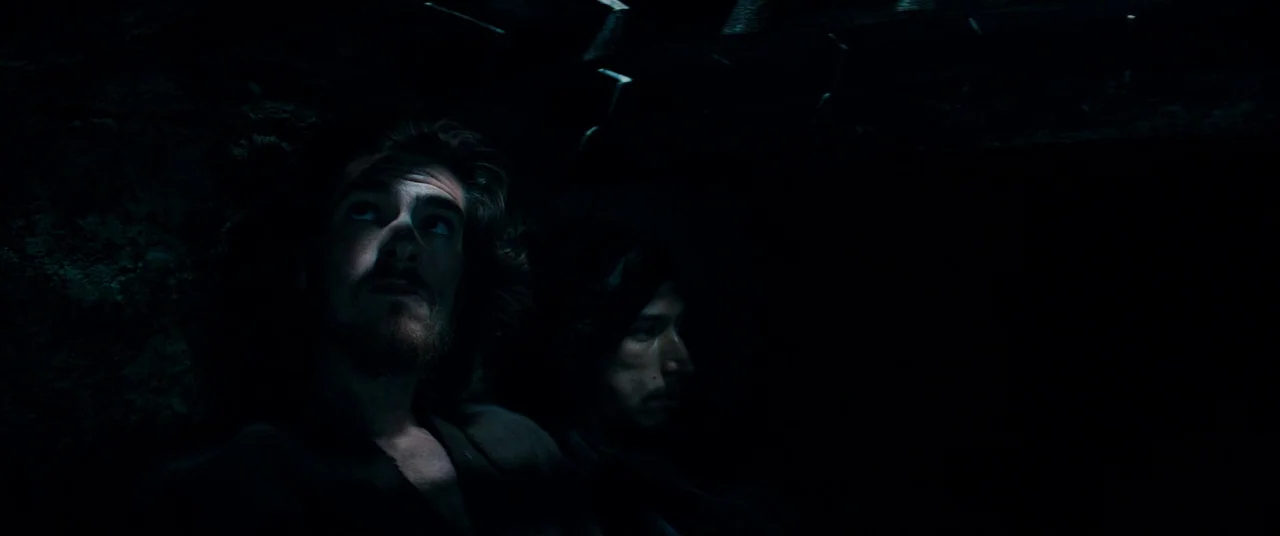 Silence (2016) movie still: medium — Two men sit in a dark, confined space with a rough, uneven texture behind them. They gaze upwards into the…