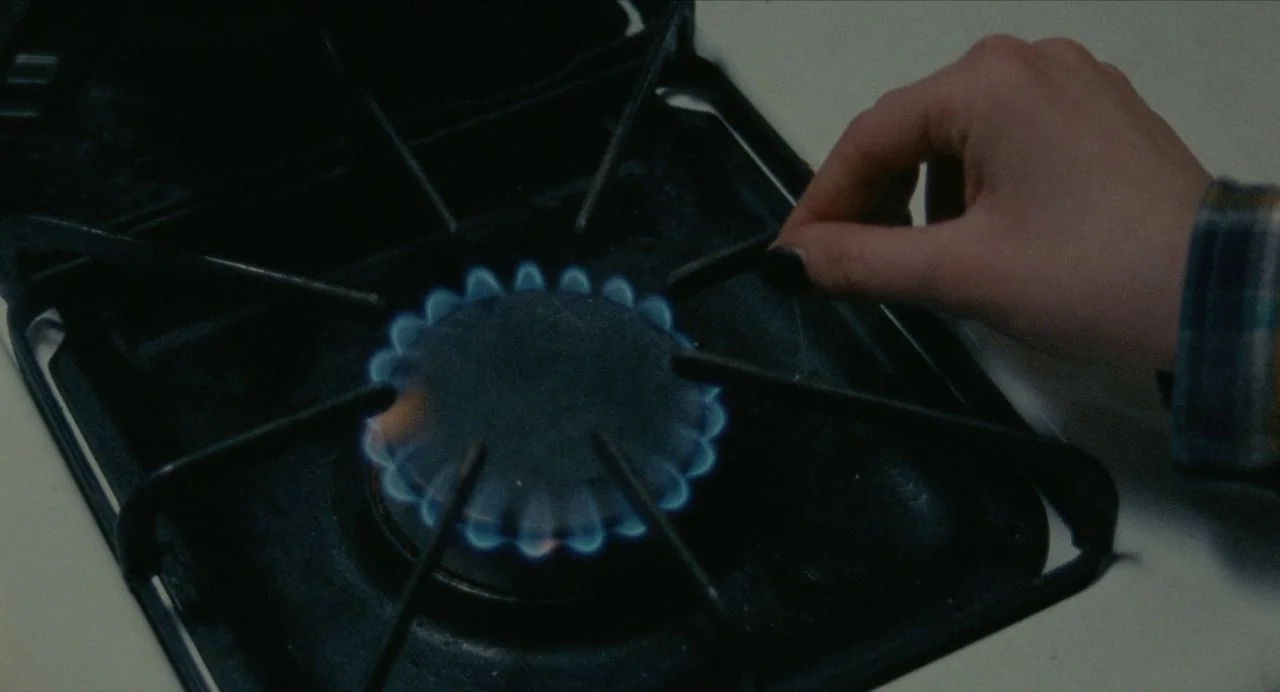 Never Rarely Sometimes Always (2020) movie still: closeup — A close view of a black metal gas stovetop with one active burner. A person's hand…