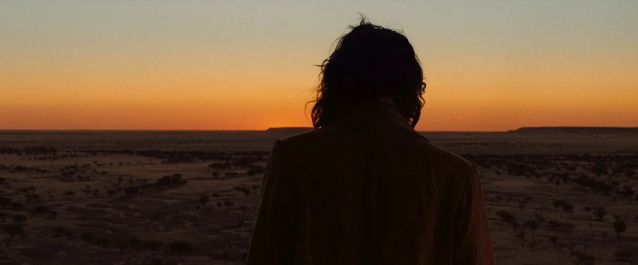 The Proposition (2005) movie still: medium — An expansive arid desert stretches toward a flat horizon under a sunset sky. A man with shoulder-length…