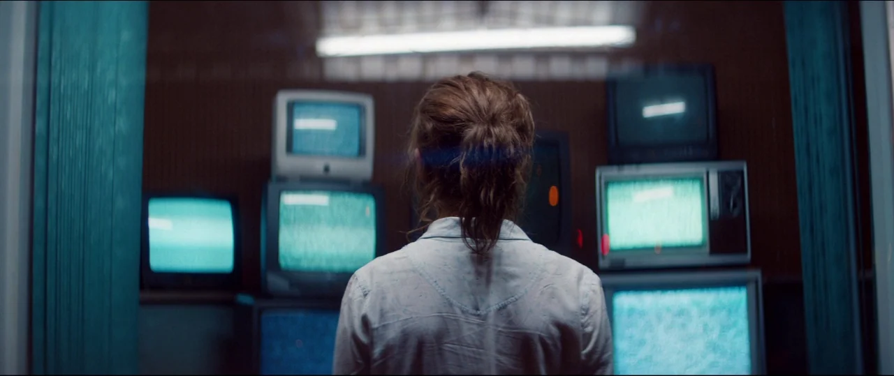 One & Two (2015) movie still: medium — A dark, cramped room contains several vintage television monitors stacked on wooden shelving units. A woman…