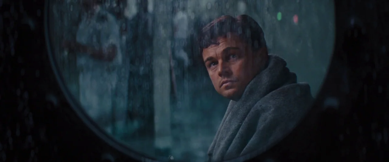 The Wolf of Wall Street (2013) movie still: closeup — A dark ship interior view through a round porthole with heavy rain on glass. A man sits by the…