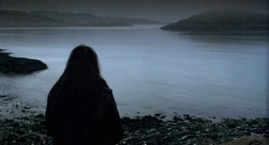 Morvern Callar (2002) movie still: medium — A dark rocky shoreline sits beside a wide, still body of water. A woman with long dark hair stands…